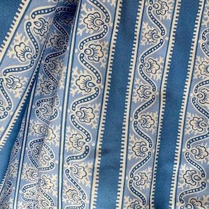 Op de afbeelding: Close-up van een blauw en wit patroonstof. Het ontwerp heeft verticale strepen met afwisselende patronen van effen blauwe en witte bloemmotieven. De stof is gevouwen en toont de textuur en de drapering.