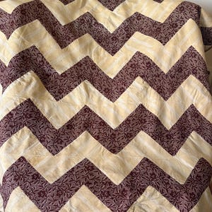 May include: A handmade quilt with a repeating chevron pattern. The quilt features alternating bands of cream-colored fabric and dark burgundy fabric with a floral print. The quilt is likely made of cotton and is suitable for a bed or sofa.