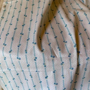 May include: Close-up of white fabric with a repeating pattern of blue vertical stripes and small blue floral accents. The fabric also features tiny grey polka dots, creating a delicate and vintage aesthetic. The fabric is folded, showing the texture.