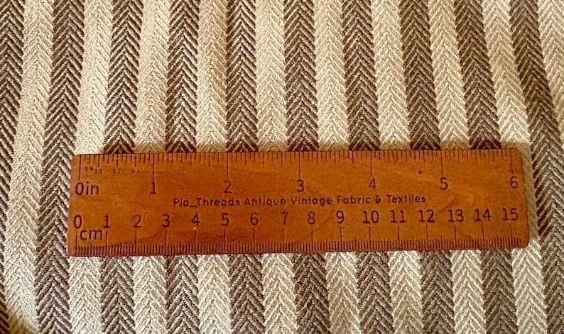 K&ouml;nnte beinhalten: Nahaufnahme eines Stoffes mit braunem und cremefarbenem Fischgr&auml;tmuster. Ein Holzlineal, in Zoll und Zentimetern markiert, liegt auf dem Stoff. Das Lineal tr&auml;gt die Aufschrift "Pio Threads Antique Vintage Fabric & Textiles."