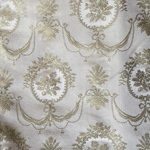 May include: A close-up of a white fabric with a repeating pattern of gold floral wreaths. The fabric has a subtle sheen and a delicate texture.