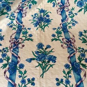 以下が含まれることがあります： 白地に花柄の繰り返し模様。青い花、緑の葉、紫のリボンと蝶結びが付いた青い縦縞模様のデザインです。生地は質感のある外観をしています。