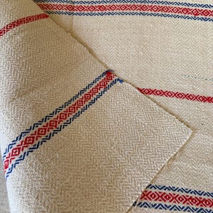 May include: Cream-colored woven fabric with red and blue geometric stripes. The fabric has a subtle herringbone weave pattern. The stripes feature a repeating diamond design in red and blue, adding a decorative element to the textile.