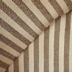 K&ouml;nnte beinhalten: Nahaufnahme eines strukturierten Stoffes mit sich wiederholendem Fischgr&auml;tmuster. Der Stoff weist abwechselnd vertikale Streifen in Beige und Dunkelbraun auf, wodurch ein optisch ansprechendes Design entsteht. Der Stoff scheint Polstermaterial zu sein.