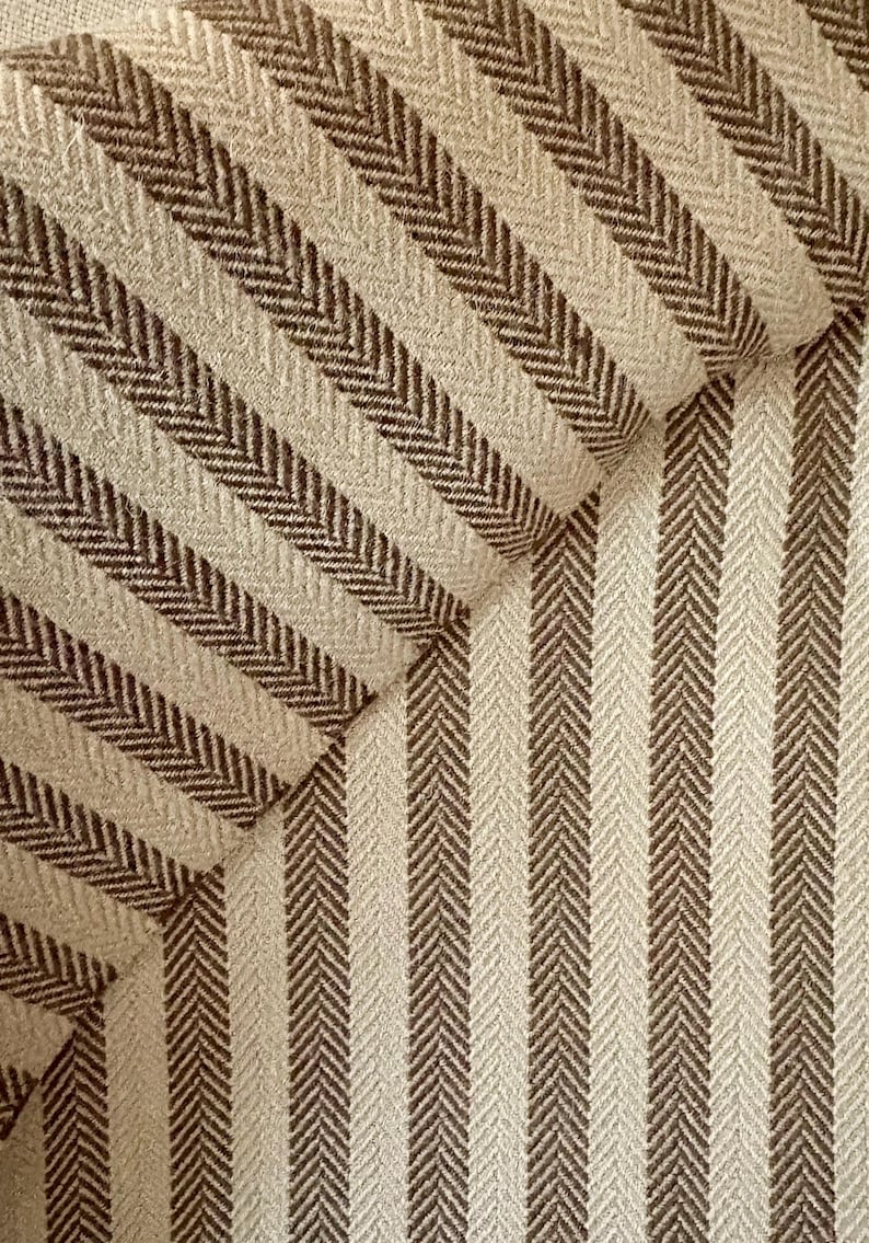 K&ouml;nnte beinhalten: Nahaufnahme eines strukturierten Stoffes mit sich wiederholendem Fischgr&auml;tmuster. Der Stoff weist abwechselnd vertikale Streifen in Beige und Dunkelbraun auf, wodurch ein optisch ansprechendes Design entsteht. Der Stoff scheint Polstermaterial zu sein.