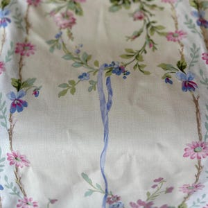 May include: Cream fabric with a floral design of pink roses, blue and pink wildflowers, and green leaves. The pattern includes vertical floral borders and a central blue ribbon, giving a delicate, vintage appearance.