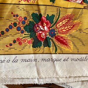 May include: Yellow fabric with a floral design featuring red, blue, and green flowers, and wheat stalks. The fabric has horizontal stripes and text in French: "Frelle, imprim&eacute; &agrave; la main, marque et mod&egrave;le d&eacute;pos&eacute;s &copy; MCMLX".
