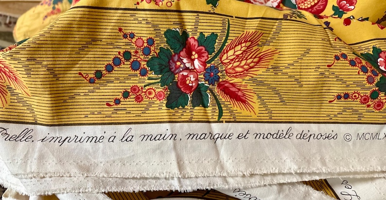 May include: Yellow fabric with a floral design featuring red, blue, and green flowers, and wheat stalks. The fabric has horizontal stripes and text in French: "Frelle, imprim&eacute; &agrave; la main, marque et mod&egrave;le d&eacute;pos&eacute;s &copy; MCMLX".