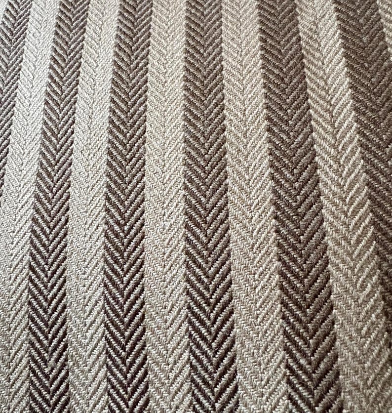 K&ouml;nnte beinhalten: Nahaufnahme eines strukturierten Stoffes mit vertikalem Streifenmuster. Die Streifen wechseln sich zwischen hellbeige und dunkelbraun ab und erzeugen ein Fischgr&auml;tmuster. Der Stoff scheint gewebt zu sein, mit einem dezenten Glanz.