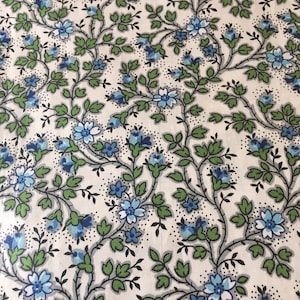 Op de afbeelding: Een witte stof met een herhalend patroon van blauwe bloemen en groene bladeren. Het patroon is een delicaat bloemenpatroon.