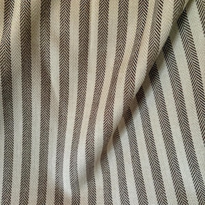 K&ouml;nnte beinhalten: Nahaufnahme eines Stoffes mit vertikalem Streifenmuster. Die Streifen wechseln sich zwischen einem hellen Beige und einem dunkleren Braungrau ab. Der Stoff hat eine subtile Fischgr&auml;twebstruktur, und die Falten erzeugen Schatten.