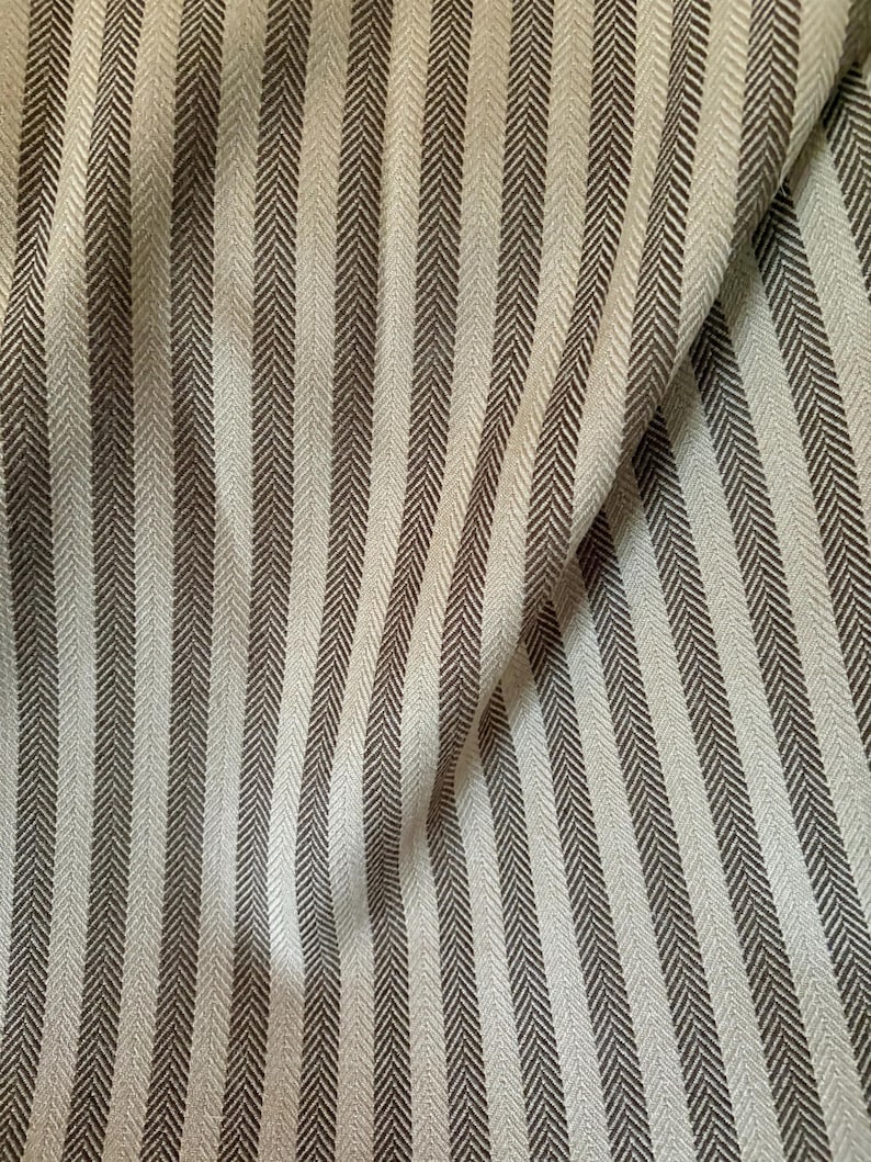 K&ouml;nnte beinhalten: Nahaufnahme eines Stoffes mit vertikalem Streifenmuster. Die Streifen wechseln sich zwischen einem hellen Beige und einem dunkleren Braungrau ab. Der Stoff hat eine subtile Fischgr&auml;twebstruktur, und die Falten erzeugen Schatten.
