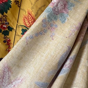 May include: Close-up of two fabrics with floral patterns. One fabric is mustard yellow with red and green flowers. The other is a light yellow fabric with a repeating pattern of pink, blue, and purple flowers and wheat stalks.