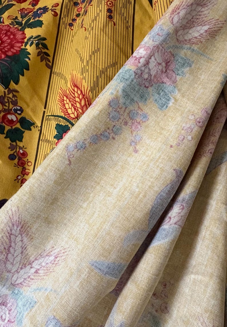 May include: Close-up of two fabrics with floral patterns. One fabric is mustard yellow with red and green flowers. The other is a light yellow fabric with a repeating pattern of pink, blue, and purple flowers and wheat stalks.