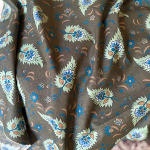 May include: Close-up of a fabric with a repeating floral pattern. The design features stylized leaves and flowers in shades of blue, green, and peach against a dark olive-green background. The fabric has a slightly wrinkled texture.