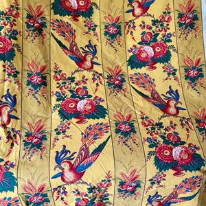May include: Yellow fabric with a repeating pattern of colorful birds, flowers, and foliage. The birds have red, blue, and teal feathers. The flowers are red and pink. The background has vertical stripes.