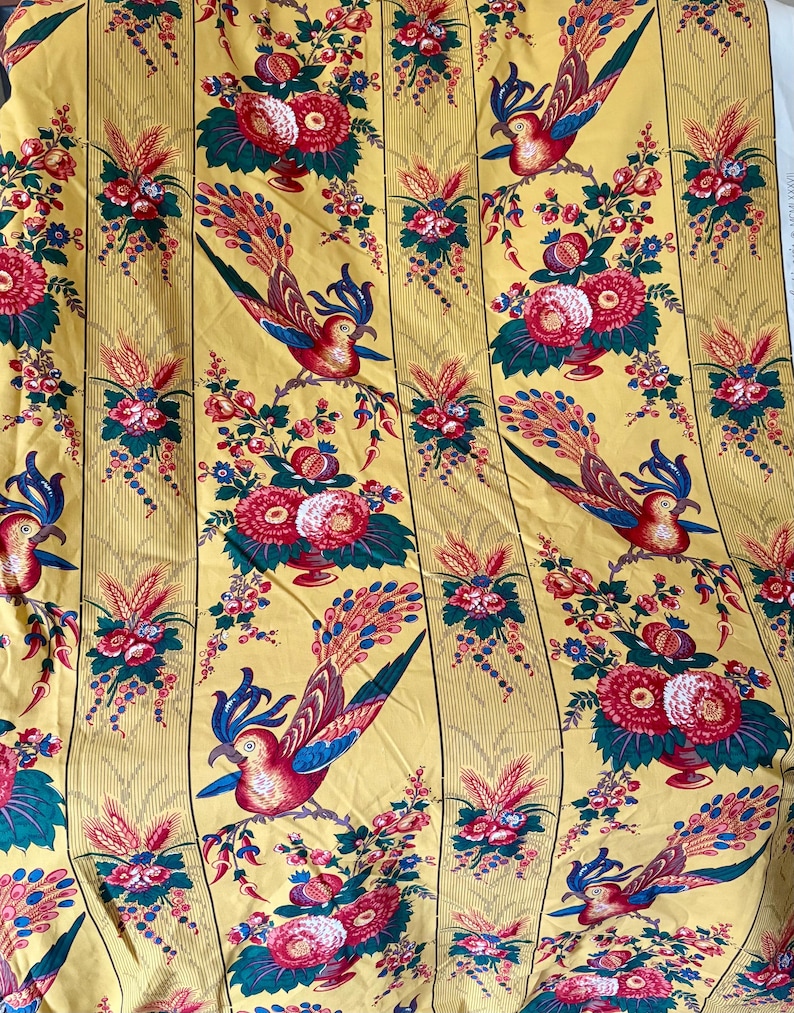 May include: Yellow fabric with a repeating pattern of colorful birds, flowers, and foliage. The birds have red, blue, and teal feathers. The flowers are red and pink. The background has vertical stripes.