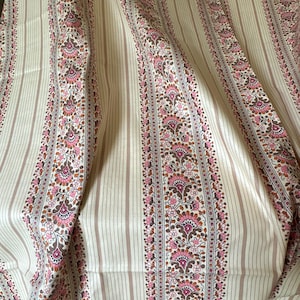 Puede incluir: Una tela con rayas verticales de anchos alternos en crema y marrón claro. Las franjas más anchas están decoradas con un patrón floral repetitivo en tonos rosa, marrón oscuro y crema, creando un diseño estructurado y repetitivo.