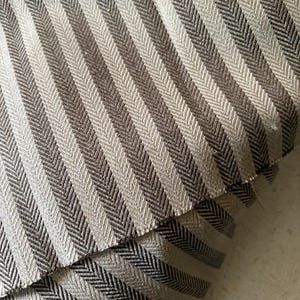 K&ouml;nnte beinhalten: Nahaufnahme eines Stoffes mit Fischgr&auml;tenmuster in Grau- und Braunt&ouml;nen. Der Stoff ist gefaltet und zeigt die Textur und das Gewebe des Materials. Die diagonalen Streifen erzeugen eine visuelle Textur.