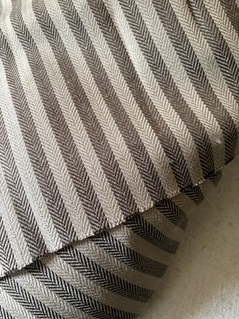 K&ouml;nnte beinhalten: Nahaufnahme eines Stoffes mit Fischgr&auml;tenmuster in Grau- und Braunt&ouml;nen. Der Stoff ist gefaltet und zeigt die Textur und das Gewebe des Materials. Die diagonalen Streifen erzeugen eine visuelle Textur.