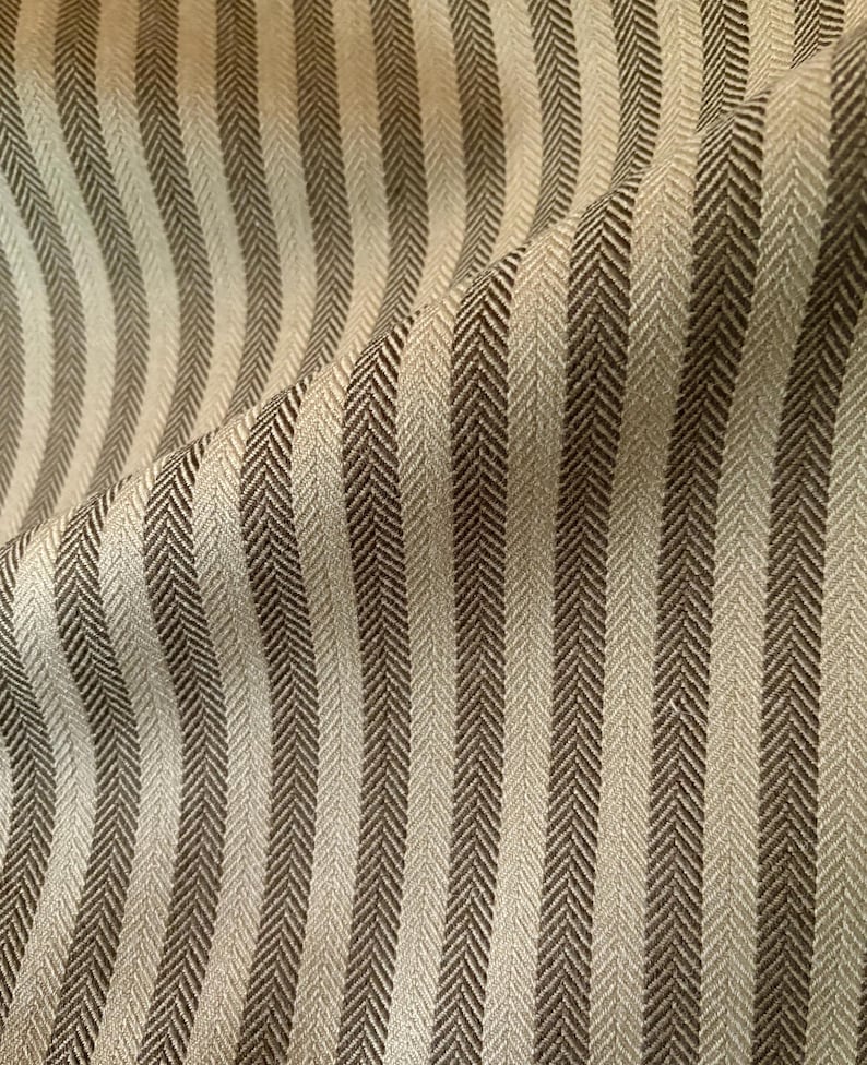 K&ouml;nnte beinhalten: Nahaufnahme eines Stoffes mit vertikalem Streifenmuster. Die Streifen wechseln sich zwischen einem hellen Beige und einem dunkleren Braun ab und erzeugen ein strukturiertes Fischgr&auml;tgewebe. Der Stoff ist leicht zerknittert und verleiht dem Design Tiefe.