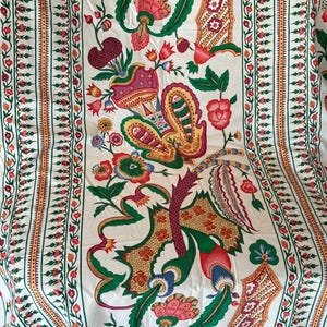 Könnte beinhalten: Ein weißer Stoff mit einem sich wiederholenden vertikalen Muster aus bunten Blumen- und Paisley-Motiven in Rot-, Grün-, Gelb- und Blautönen. Das Design umfasst komplizierte Ränder mit kleinen roten Blütenknospen und grünen Blättern.