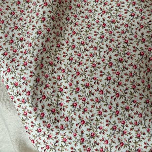 May include: Close-up of a white fabric covered in a pattern of small red roses and green leaves. The floral design is densely packed, creating a textured appearance. The fabric is slightly wrinkled.