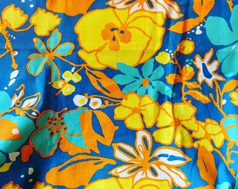 Op de afbeelding: Een blauwe stof met een bloemenpatroon in tinten geel, oranje en turquoise. De bloemen zijn gestileerd en hebben een retro-uitstraling.