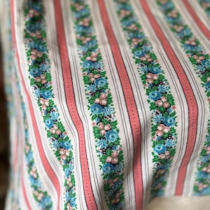 May include: Close-up of a fabric with a repeating pattern of vertical stripes. Alternating stripes are white, coral pink, and black. The white stripes feature a floral design with blue and pink flowers and green leaves. The fabric appears to be cotton.