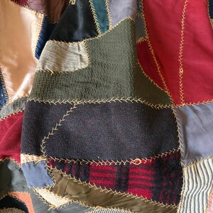 May include: Close-up of a patchwork quilt with various fabric textures and colors. The quilt features squares of red, black, green, blue, and patterned fabrics, stitched together with gold thread. The quilt's design is a mix of textures and patterns.