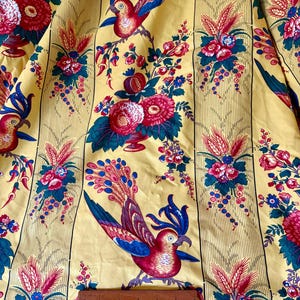 May include: Yellow fabric with a repeating pattern of red, blue, and pink birds, flowers, and foliage. Vertical stripes run the length of the fabric. A ruler shows measurements in inches and centimeters.