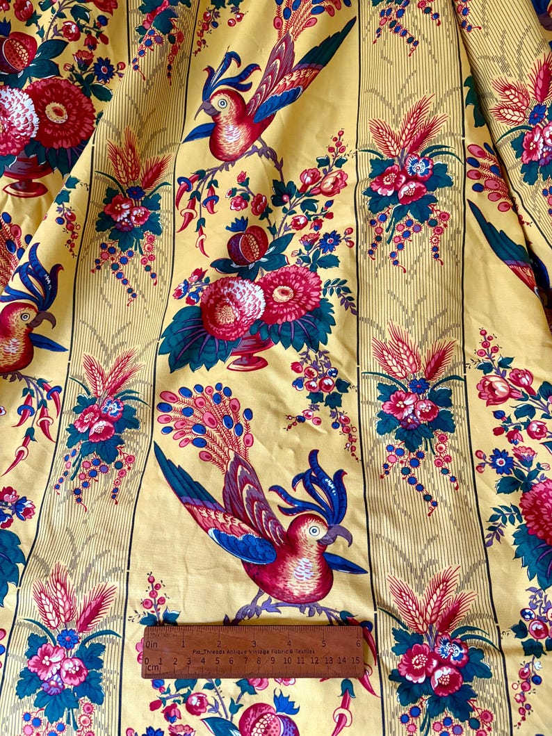 May include: Yellow fabric with a repeating pattern of red, blue, and pink birds, flowers, and foliage. Vertical stripes run the length of the fabric. A ruler shows measurements in inches and centimeters.