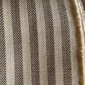 K&ouml;nnte beinhalten: Nahaufnahme eines strukturierten Stoffes mit Fischgr&auml;tmuster. Der Stoff weist vertikale Streifen in Beige- und Braunt&ouml;nen auf. Die Kanten des Stoffes sind sichtbar und zeigen ein fransiges Detail. Dieser Stoff eignet sich f&uuml;r Polster oder Heimdekoration.