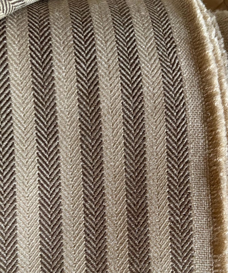 K&ouml;nnte beinhalten: Nahaufnahme eines strukturierten Stoffes mit Fischgr&auml;tmuster. Der Stoff weist vertikale Streifen in Beige- und Braunt&ouml;nen auf. Die Kanten des Stoffes sind sichtbar und zeigen ein fransiges Detail. Dieser Stoff eignet sich f&uuml;r Polster oder Heimdekoration.