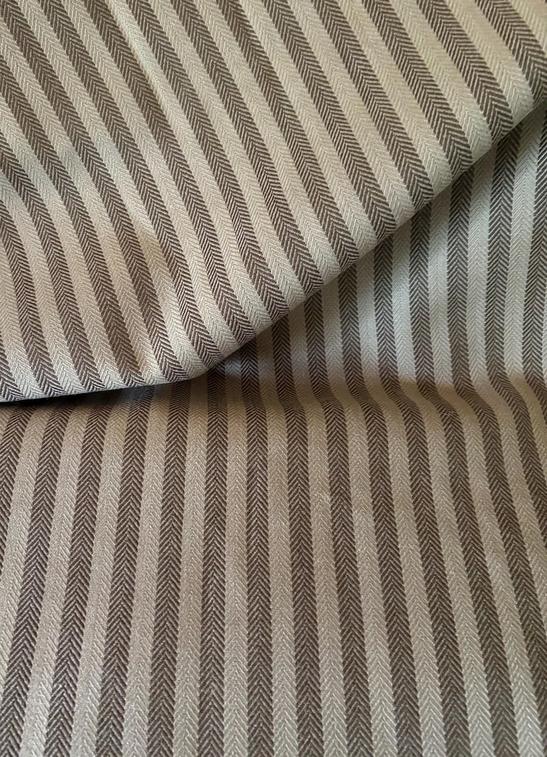 K&ouml;nnte beinhalten: Nahaufnahme eines Stoffes mit sich wiederholendem vertikalem Streifenmuster. Die Streifen wechseln sich zwischen einem hellen Beige und einem dunkleren Braun-Grau ab. Der Stoff ist gefaltet und zeigt die Textur und das Gewebe des Materials.