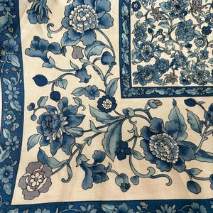 Può includere: Primo piano di un tessuto con un motivo floreale nei toni del blu e del bianco. Il design presenta grandi fiori stilizzati e foglie, con un bordo di motivi floreali più piccoli. L'estetica generale ricorda la tradizionale Chinoiserie.