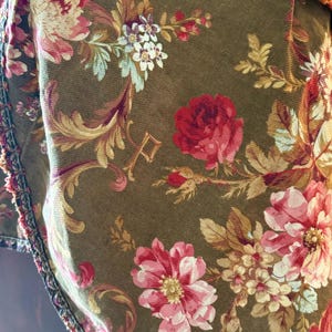 Pu&ograve; includere: Primo piano di un tessuto marrone con un motivo floreale. Il tessuto presenta rose rosa, rosse e bianche con foglie verdi. Il tessuto ha una superficie testurizzata e un bordo a frangia.