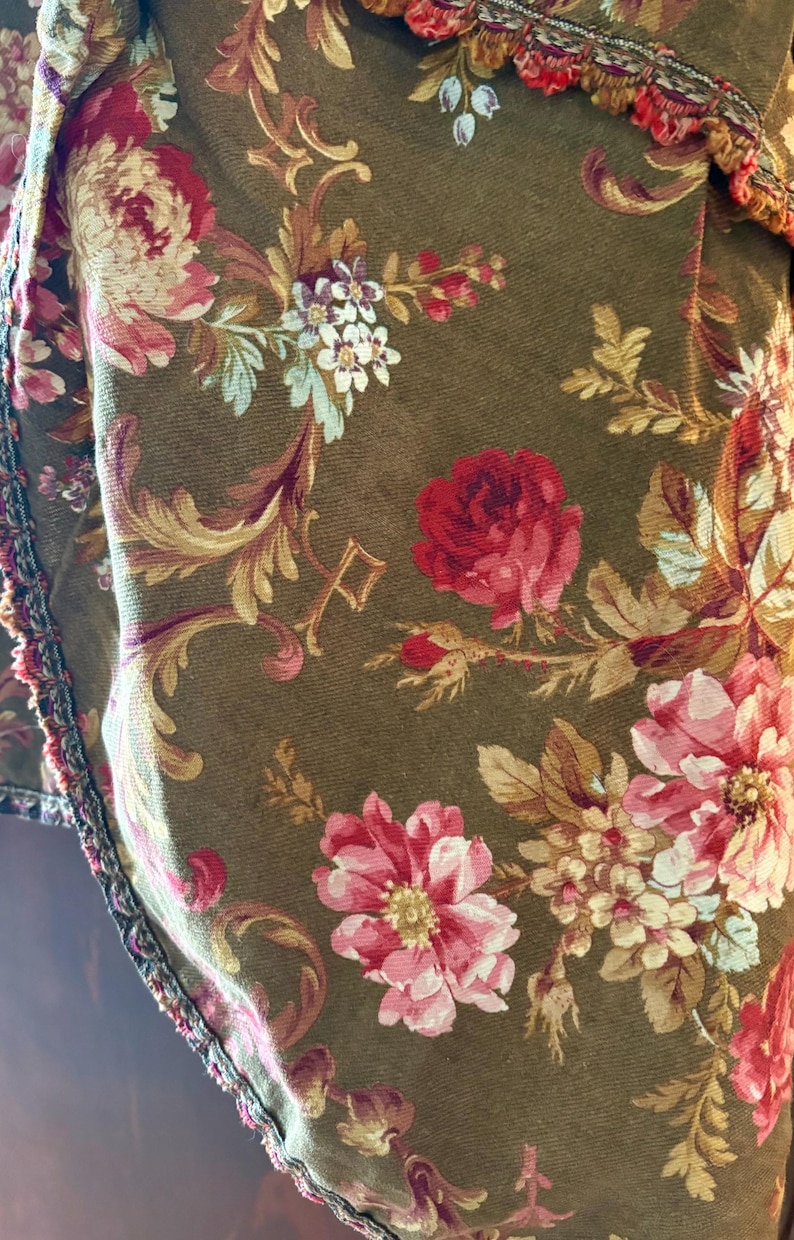 Pu&ograve; includere: Primo piano di un tessuto marrone con un motivo floreale. Il tessuto presenta rose rosa, rosse e bianche con foglie verdi. Il tessuto ha una superficie testurizzata e un bordo a frangia.