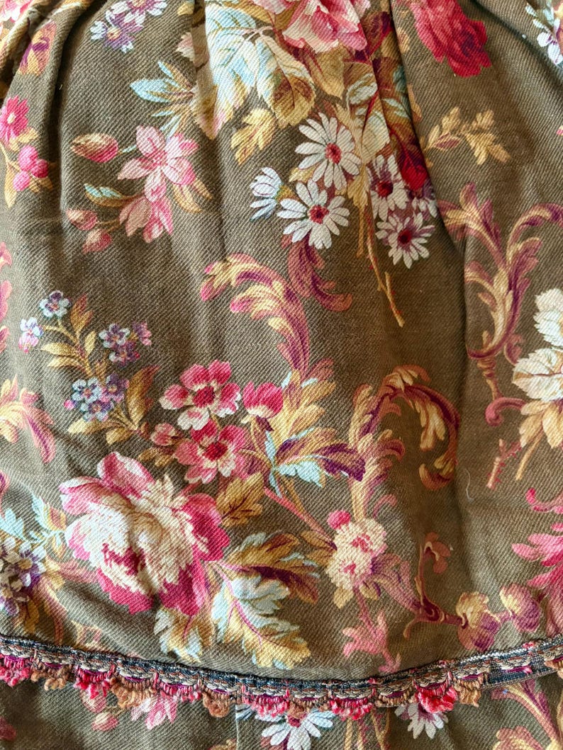 Pu&ograve; includere: Un primo piano di un tessuto marrone con un motivo floreale. Il tessuto presenta un motivo floreale rosa, bianco e giallo su sfondo verde. Il tessuto ha un bordo con una frangia.