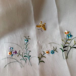 以下が含まれることがあります： 黄色、緑、青、紫の色合いの刺繍された花と蝶が描かれた白い布地です。