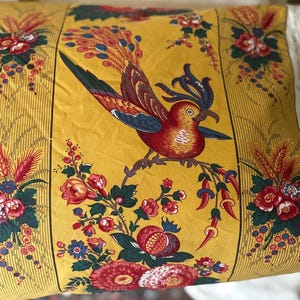 May include: A decorative pillow with a yellow background featuring a detailed bird and floral design. The bird is red, brown, and blue, perched on a branch with red and pink flowers. The pillow has a vertical striped border.