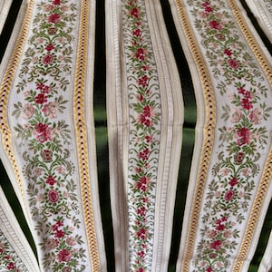 May include: Close-up of a fabric with a repeating pattern of vertical stripes. Alternating stripes are white with floral embroidery and dark green. The floral embroidery features red and pink flowers with green leaves. Gold trim borders the floral stripes.