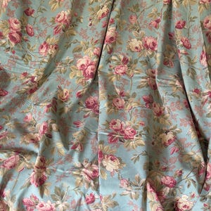 Puede incluir: Una tela de color azul claro con un patrón floral repetitivo de rosas rosas y crema con hojas y tallos verdes. El material está drapeado en pliegues suaves, mostrando el denso estampado botánico en toda la superficie del tejido.