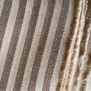 K&ouml;nnte beinhalten: Nahaufnahme eines strukturierten Stoffes mit Fischgr&auml;tenmuster. Der Stoff weist abwechselnd vertikale Streifen in Hellbeige und Dunkelbraun auf. Die Textur ist sichtbar, mit einem weichen, leicht verschwommenen Aussehen.