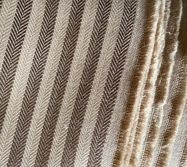 K&ouml;nnte beinhalten: Nahaufnahme eines strukturierten Stoffes mit Fischgr&auml;tenmuster. Der Stoff weist abwechselnd vertikale Streifen in Hellbeige und Dunkelbraun auf. Die Textur ist sichtbar, mit einem weichen, leicht verschwommenen Aussehen.
