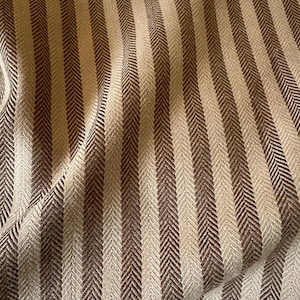 K&ouml;nnte beinhalten: Nahaufnahme eines Stoffes mit vertikalem Streifenmuster. Die Streifen wechseln sich zwischen hellbeige und dunkelbraun ab und erzeugen ein strukturiertes Fischgr&auml;tgewebe. Der Stoff ist leicht zerknittert und zeigt den weichen Fall des Materials.