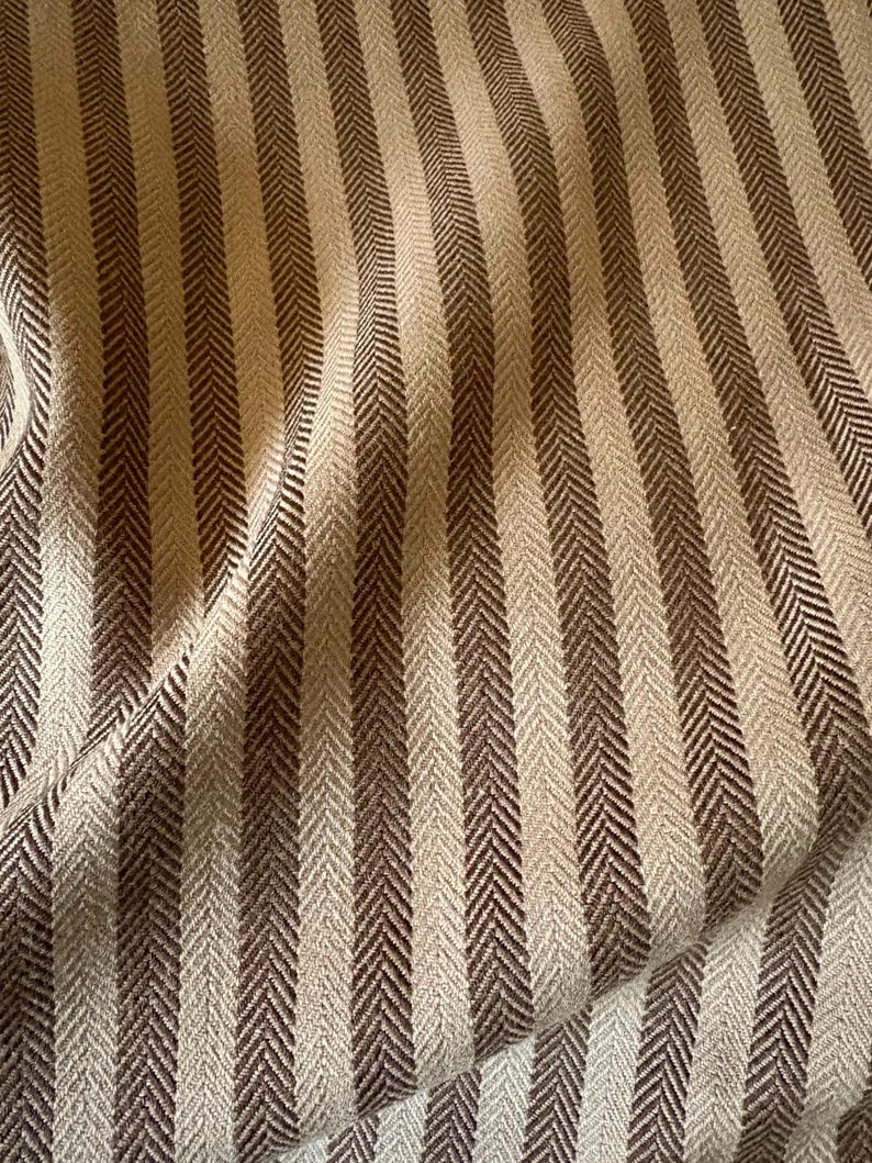 K&ouml;nnte beinhalten: Nahaufnahme eines Stoffes mit vertikalem Streifenmuster. Die Streifen wechseln sich zwischen hellbeige und dunkelbraun ab und erzeugen ein strukturiertes Fischgr&auml;tgewebe. Der Stoff ist leicht zerknittert und zeigt den weichen Fall des Materials.