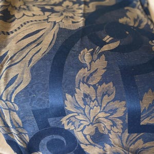 May include: Close-up of a textile with a repeating floral pattern in gold and navy blue. The design features stylized flowers, leaves, and swirling geometric shapes. The fabric has a subtle sheen, suggesting a luxurious material.