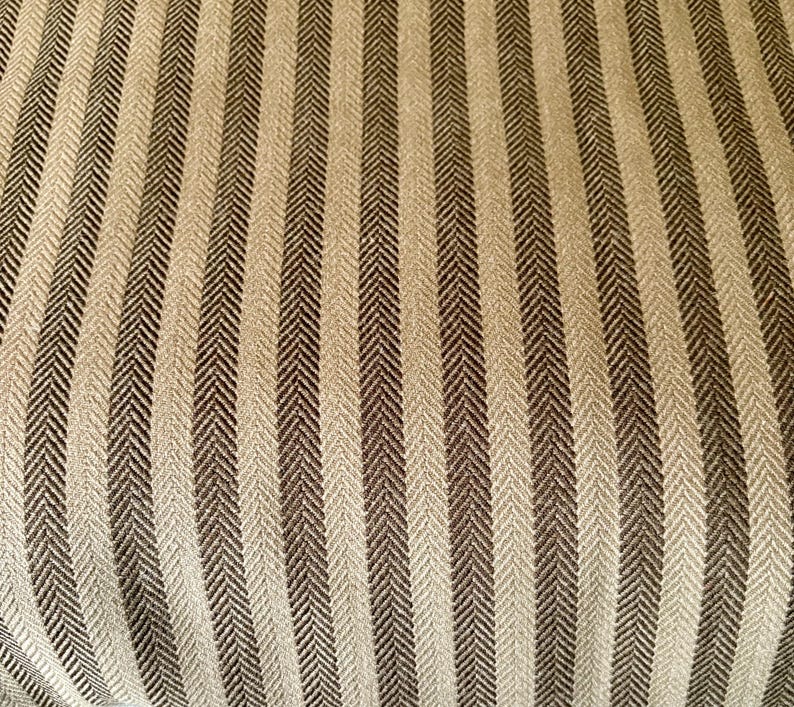 K&ouml;nnte beinhalten: Nahaufnahme eines strukturierten Stoffes mit einem sich wiederholenden vertikalen Streifenmuster. Die Streifen wechseln sich zwischen einem hellen Beige und einem dunklen Braun ab. Der Stoff hat ein subtiles Fischgr&auml;tgewebe, das dem Design Tiefe und visuelles Interesse verleiht.