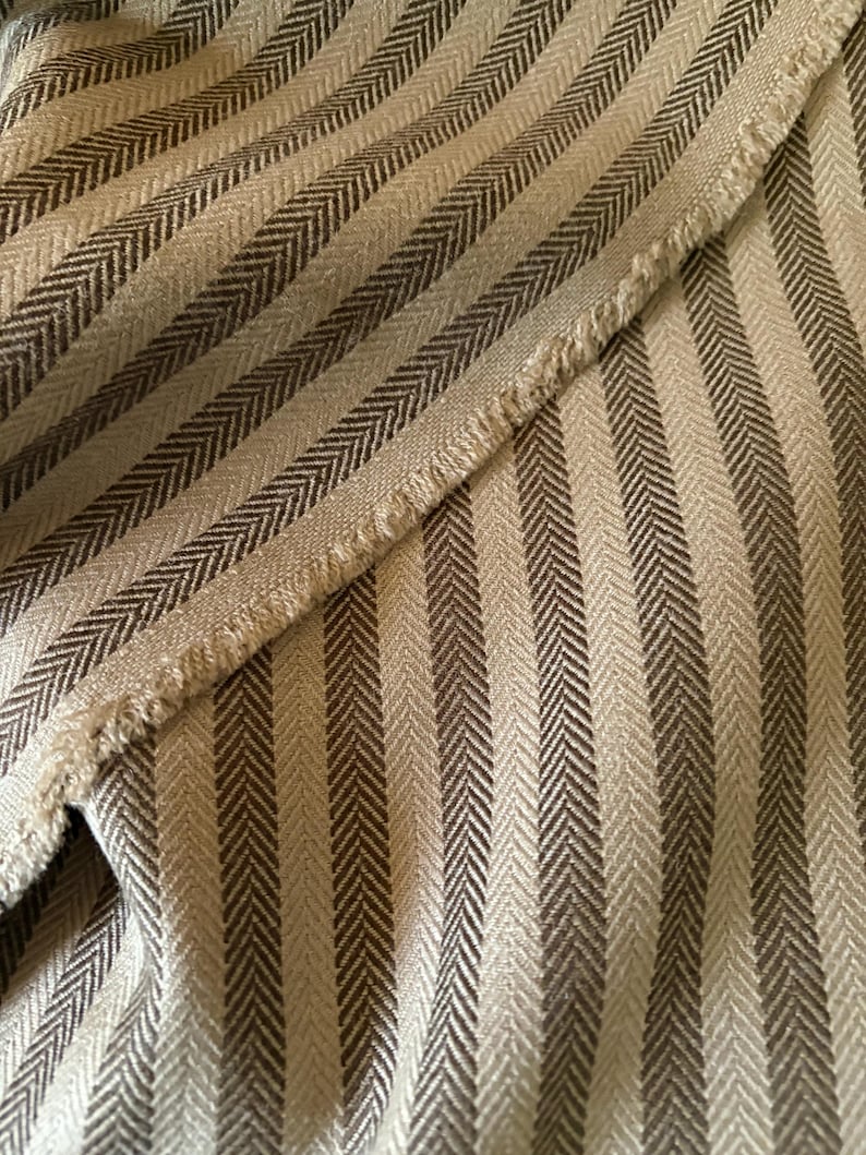 K&ouml;nnte beinhalten: Nahaufnahme eines Stoffes mit Fischgr&auml;tmuster. Der Stoff weist abwechselnd vertikale Streifen in Hellbeige und Dunkelbraun auf. Der Rand des Stoffes ist ausgefranst und gibt den Blick auf das Gewebe frei.